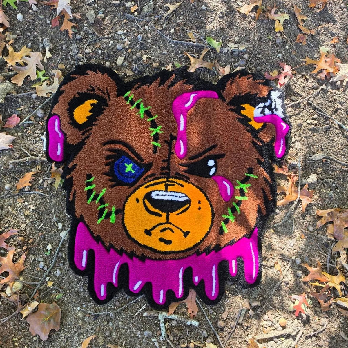 Drip bear rug with a stitched face, blue eye and pink goop, lying on autumn leaves